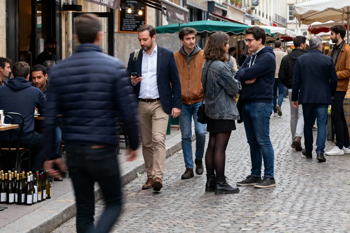 Rue animée avec passants, cafés, et bouteilles de vin. Conversations et ambiance décontractée en extérieur.