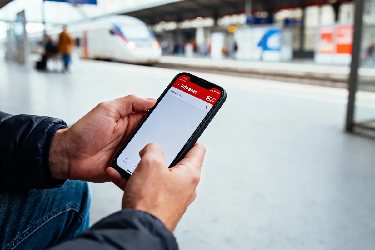Homme utilisant un smartphone avec application ouverte, en gare, devant trains à quai.