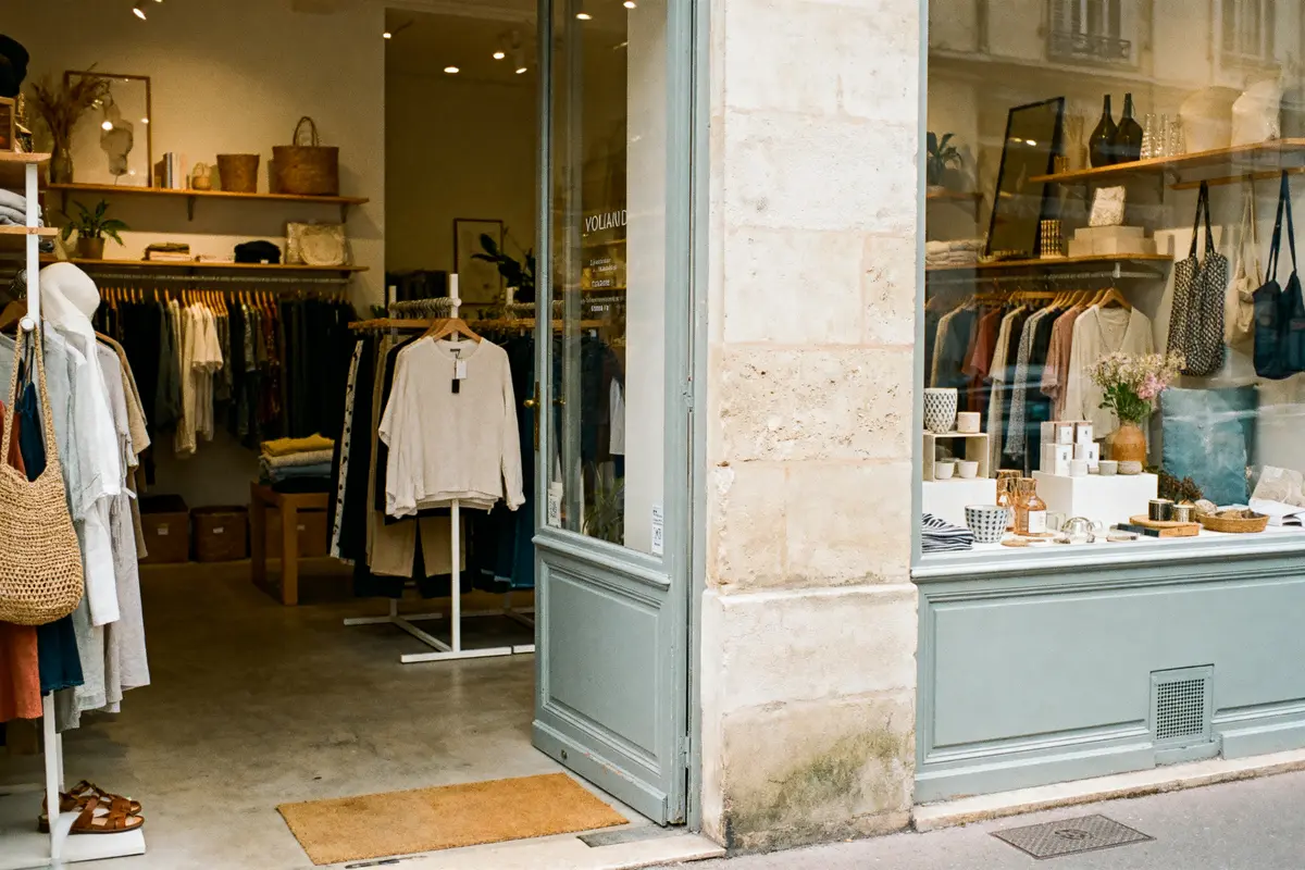 Boutique chaleureuse avec vêtements suspendus, accessoires, paniers et vaisselle, affichés autour de l'entrée vitrée accueillante.