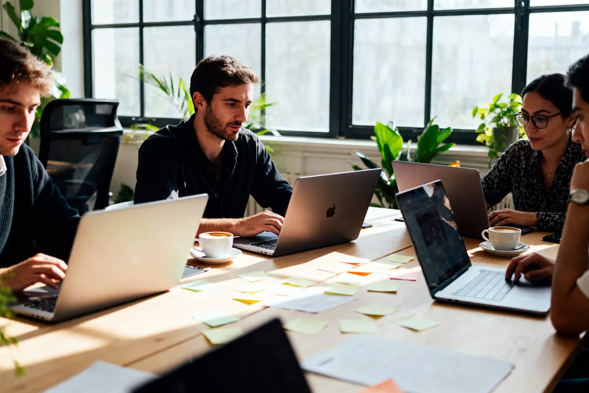 Quatre personnes travaillent sur des ordinateurs portables autour d'une table, avec des documents et des stickers.