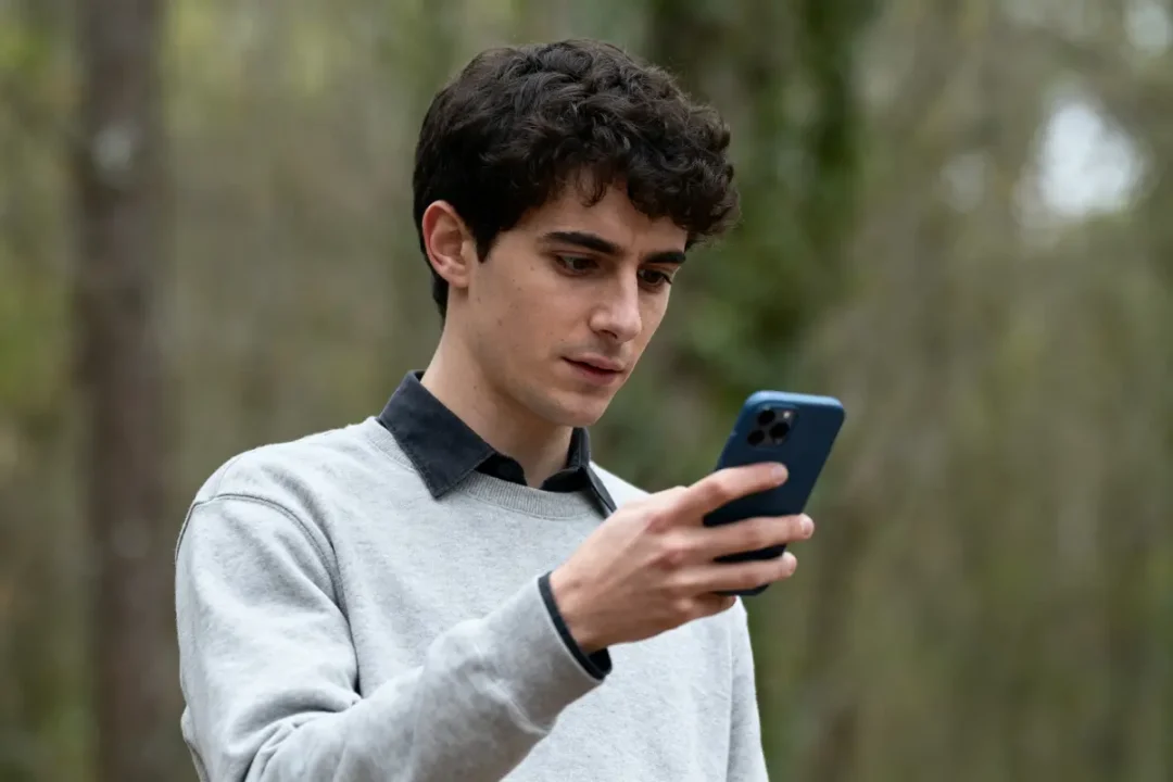 Jeune homme utilisant un smartphone en extérieur, portant un pull gris et une chemise noire.