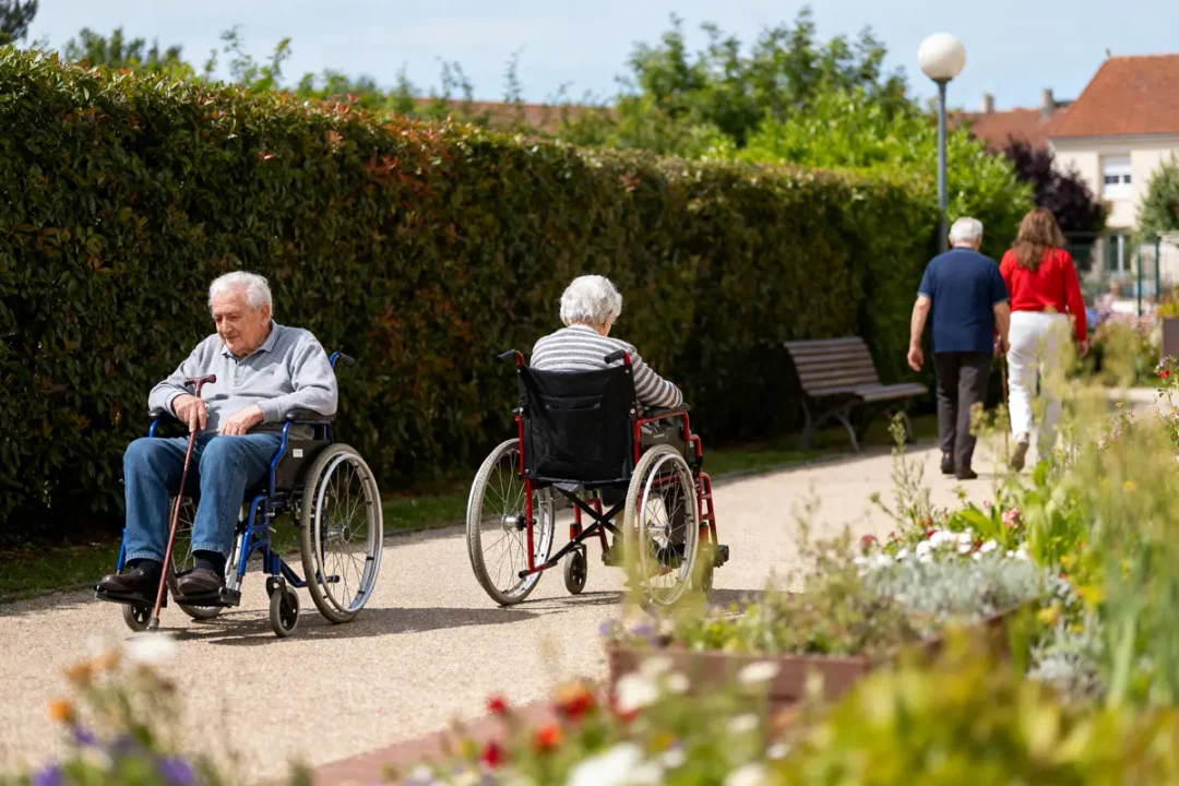 Deux personnes âgées en fauteuil roulant dans un jardin, suivies par un couple qui marche.