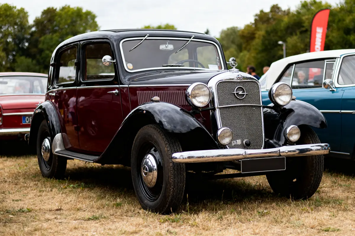Voiture classique noire et bordeaux exposée en extérieur, avec d'autres véhicules anciens en arrière-plan.