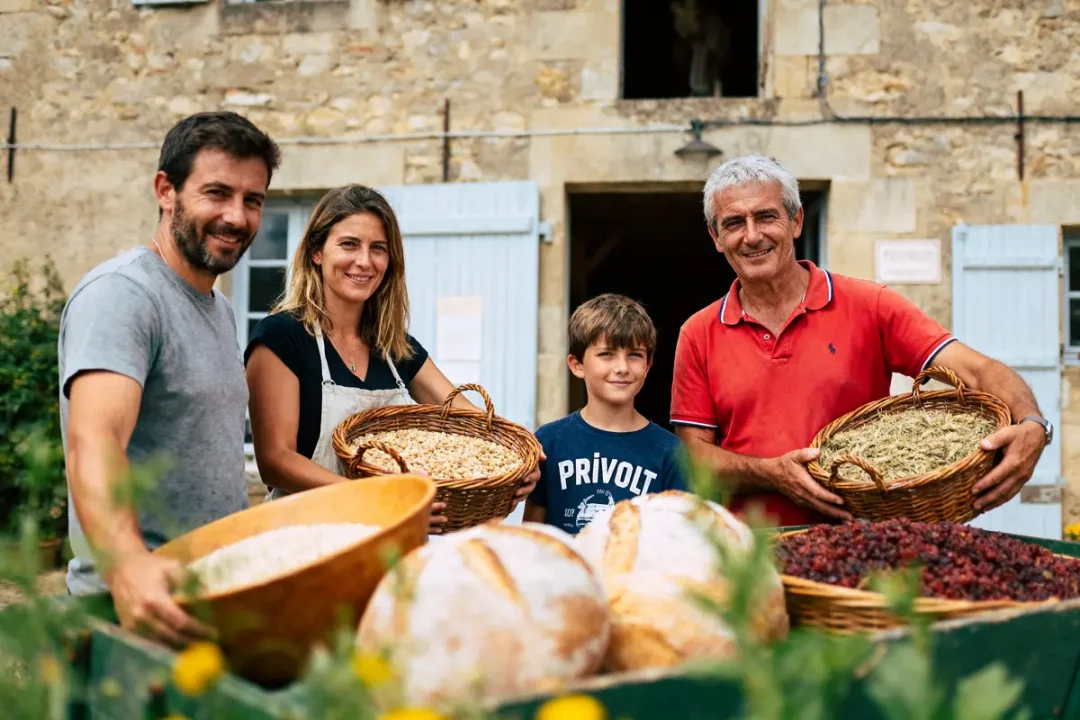 Quatre personnes souriantes avec des paniers de produits frais devant une maison en pierre.