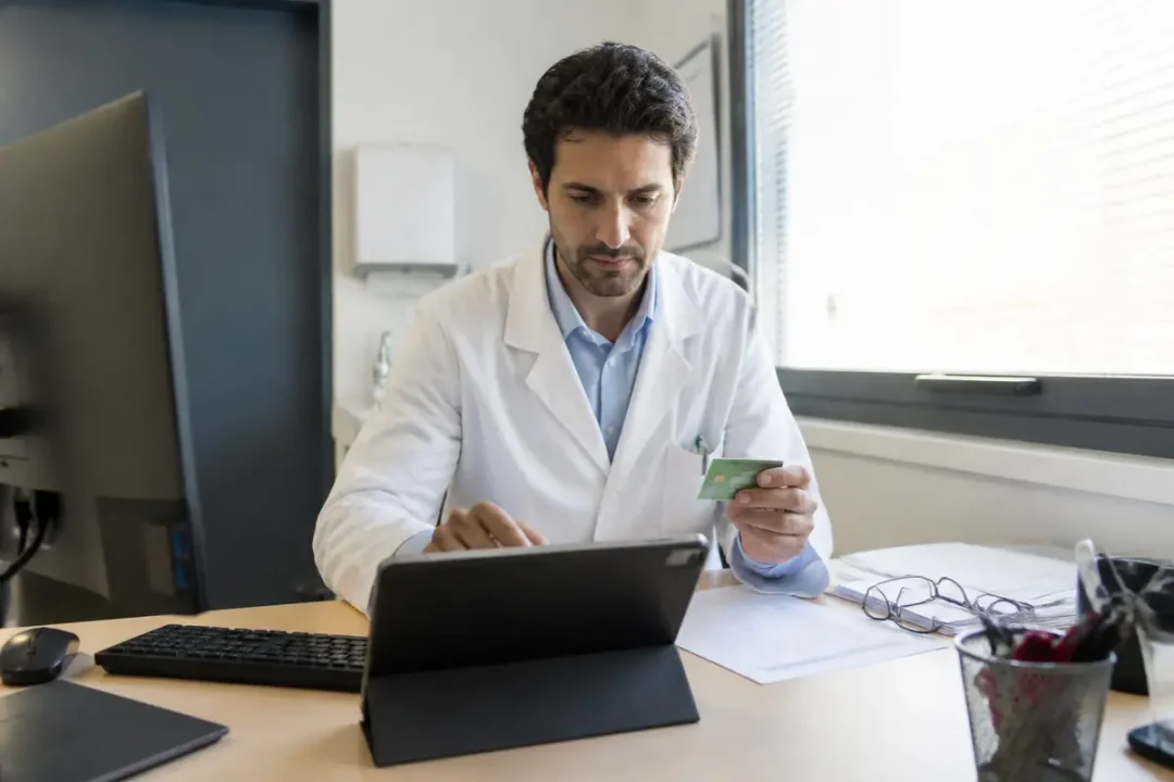 Un médecin utilisant une tablette dans un bureau, tenant une carte verte, concentré sur l'écran.