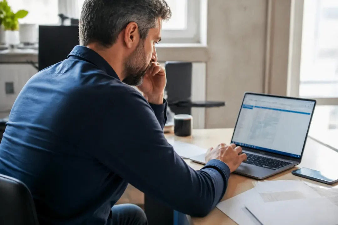 Homme travaillant sur ordinateur portable dans un bureau, concentré sur l'écran, avec téléphone et documents.