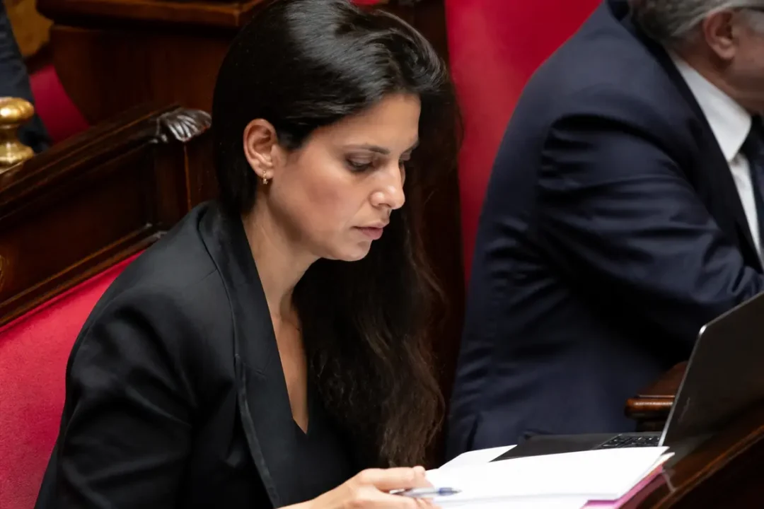 Une femme concentrée lit des documents à une table en bois; ordinateur portable ouvert à ses côtés.