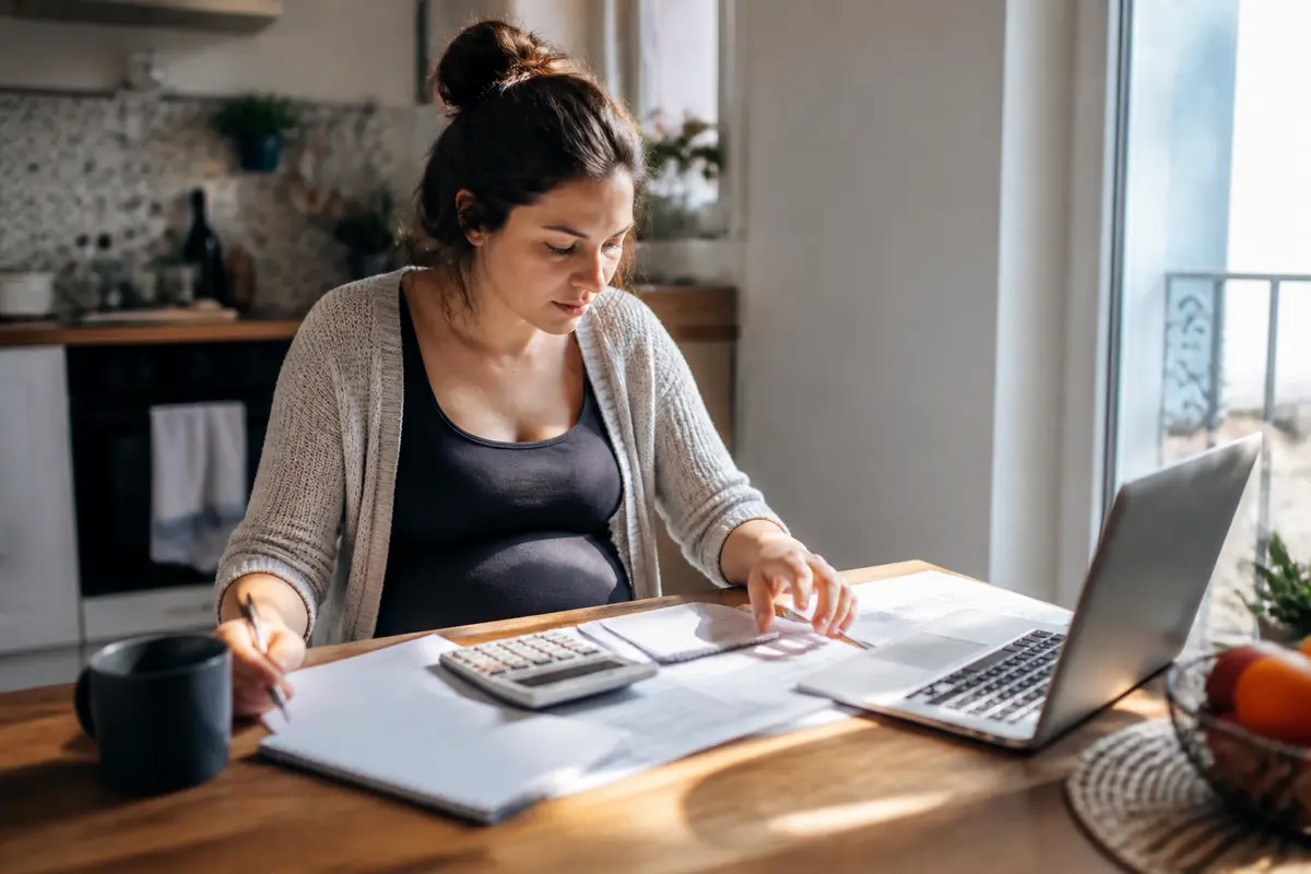 Une femme enceinte travaillant à domicile, utilisant calculatrice et ordinateur portable sur une table éclairée par le soleil.