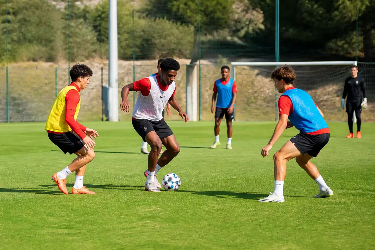 Des joueurs de football s'entraînent intensément sur un terrain ensoleillé, concentrés sur le ballon.
