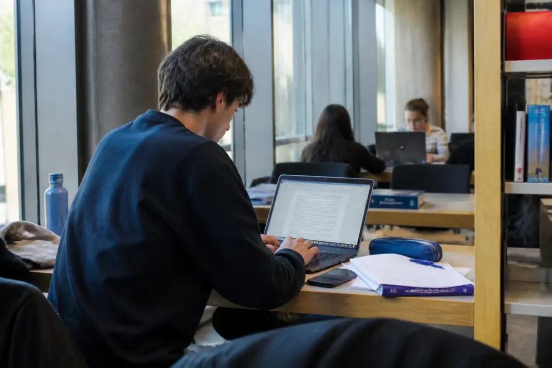 Étudiants travaillant sur ordinateurs portables dans une bibliothèque, avec livres et fournitures scolaires.