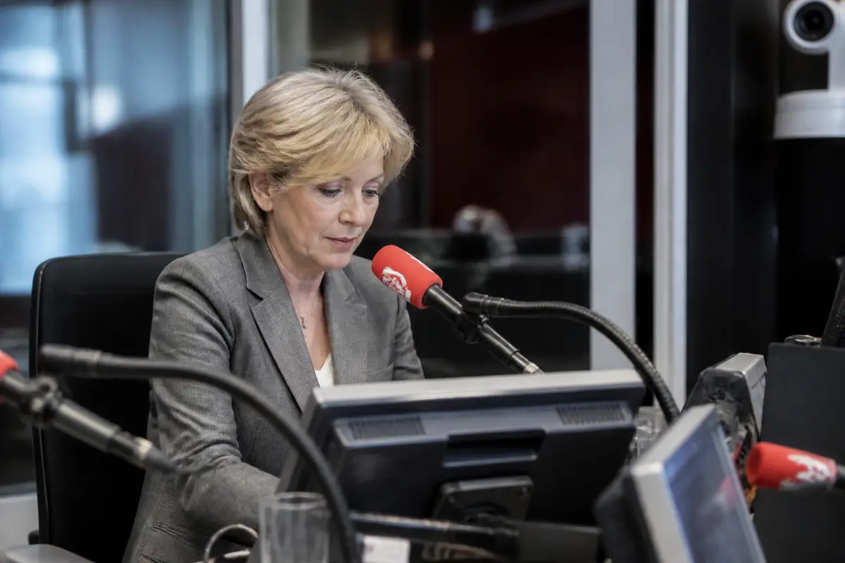 Une femme en costume gris parlant devant un micro dans un studio radio professionnel.