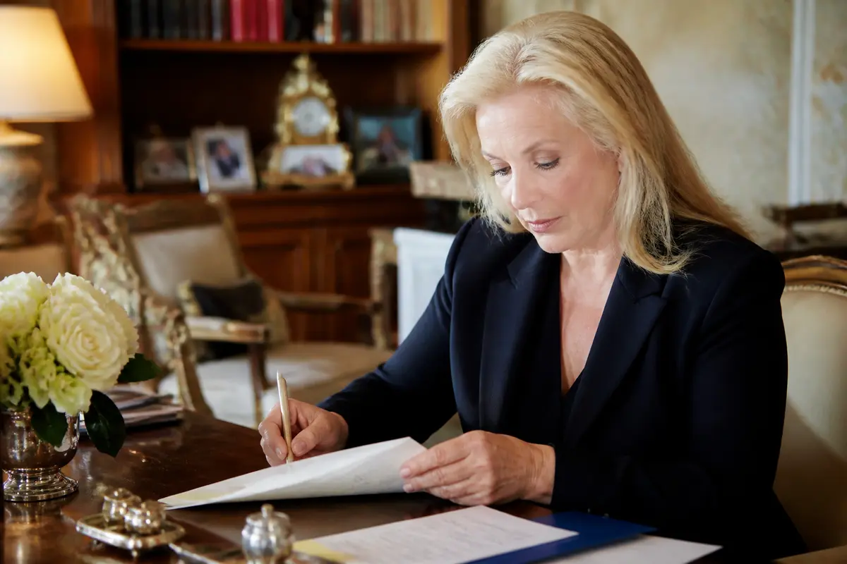 Femme blonde en costume sombre écrivant à un bureau élégant avec fleurs et horloge décorative.