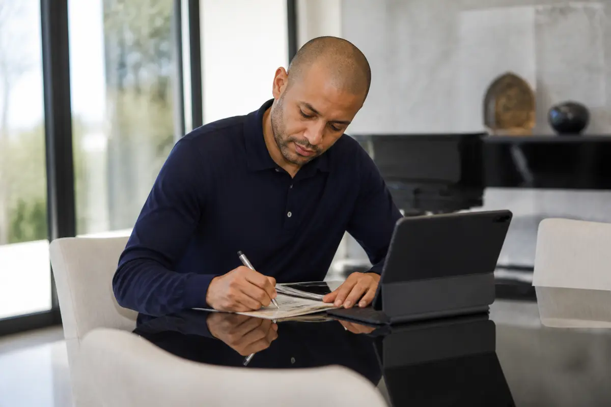 Un homme écrit sur un cahier, assis à une table avec une tablette numérique.