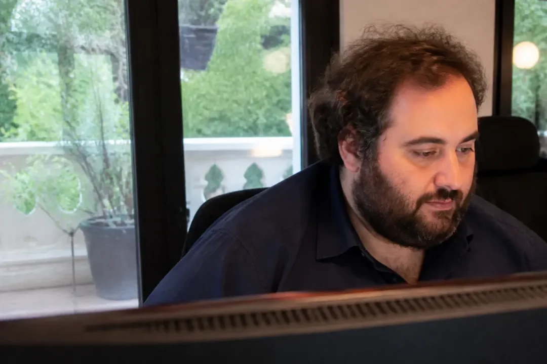 Homme barbu travaillant devant un ordinateur, regard concentré, vue sur terrasse verdoyante à travers la fenêtre.