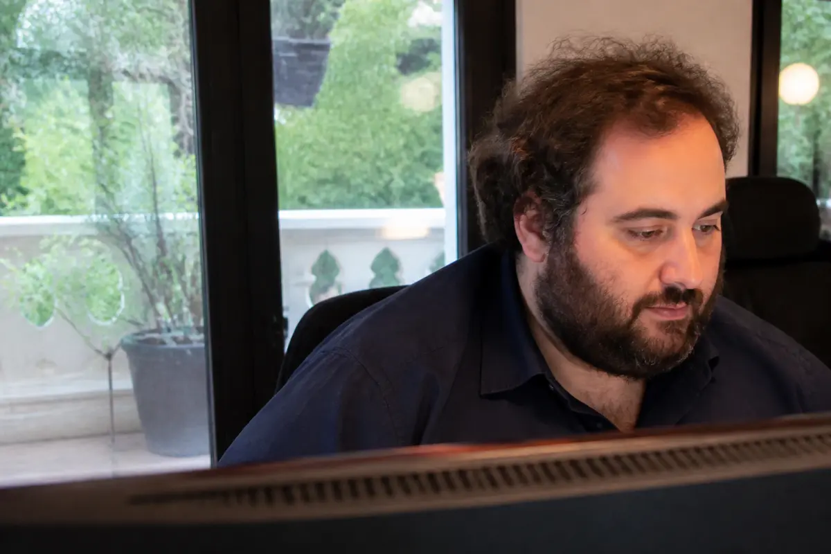 Homme barbu travaillant devant un ordinateur, regard concentré, vue sur terrasse verdoyante à travers la fenêtre.