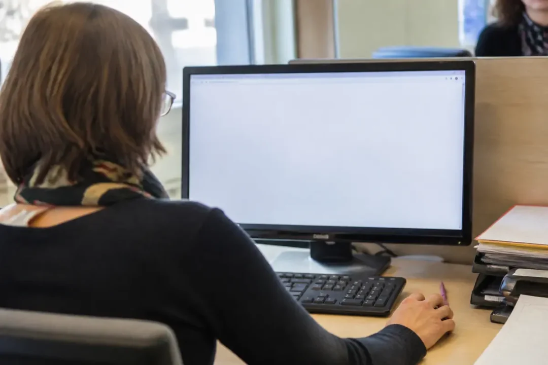 Une femme de dos travaille sur un ordinateur avec écran blanc dans un bureau.
