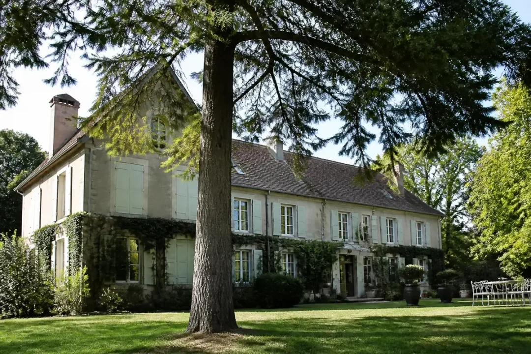Grande maison en pierre avec volets verts, entourée de verdure et d'arbres, sous ciel ensoleillé.