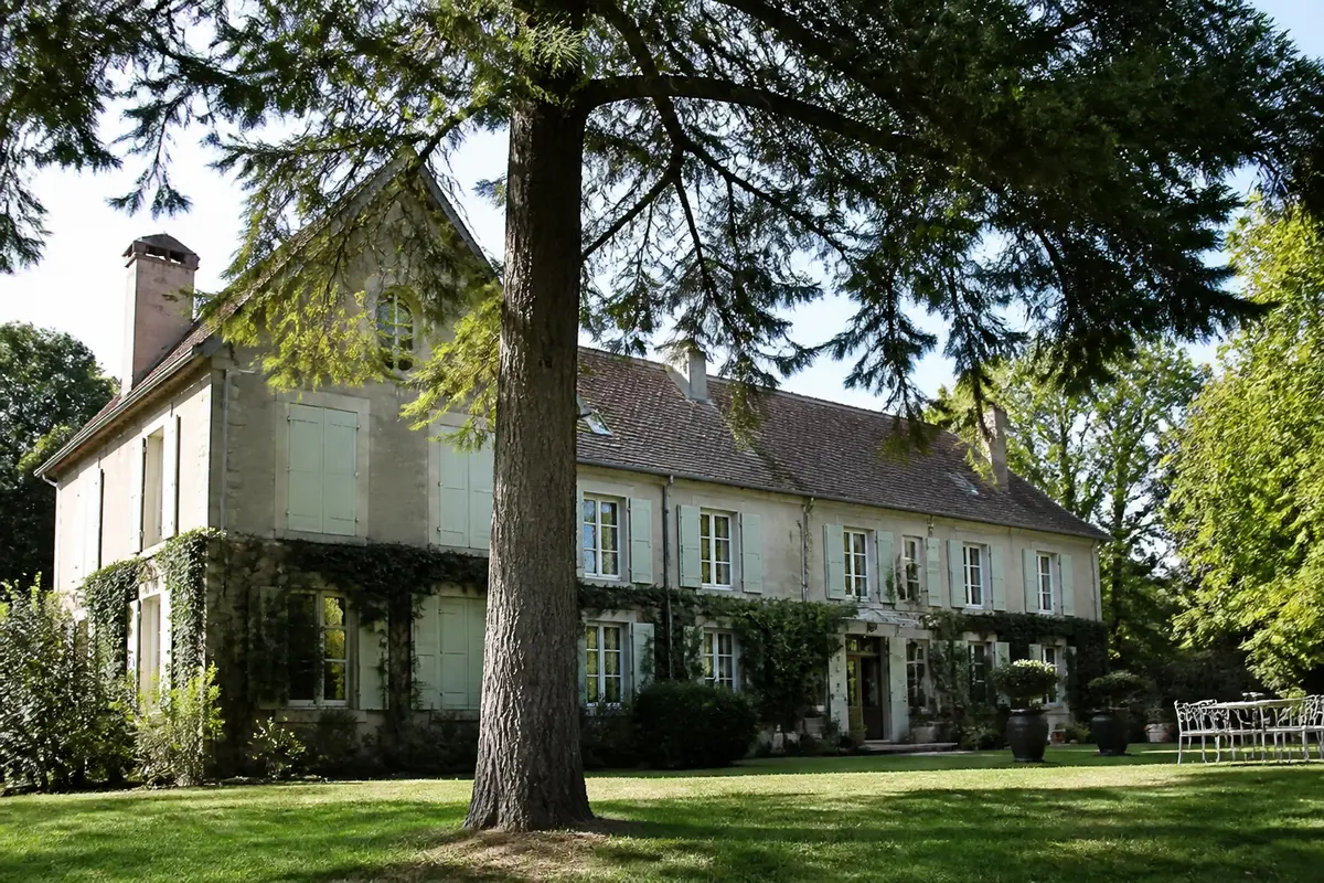 Grande maison en pierre avec volets verts, entourée de verdure et d'arbres, sous ciel ensoleillé.