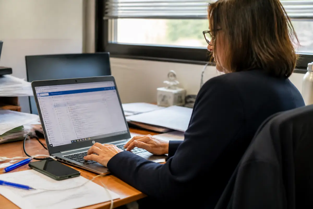 Femme travaillant sur ordinateur portable dans un bureau, entourée de documents et d'un téléphone portable.