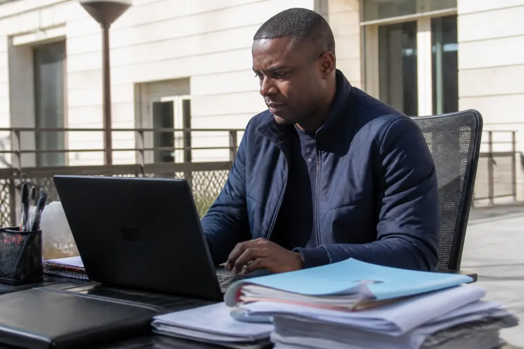 Un homme travaille sur un ordinateur portable à l'extérieur, entouré de documents empilés.
