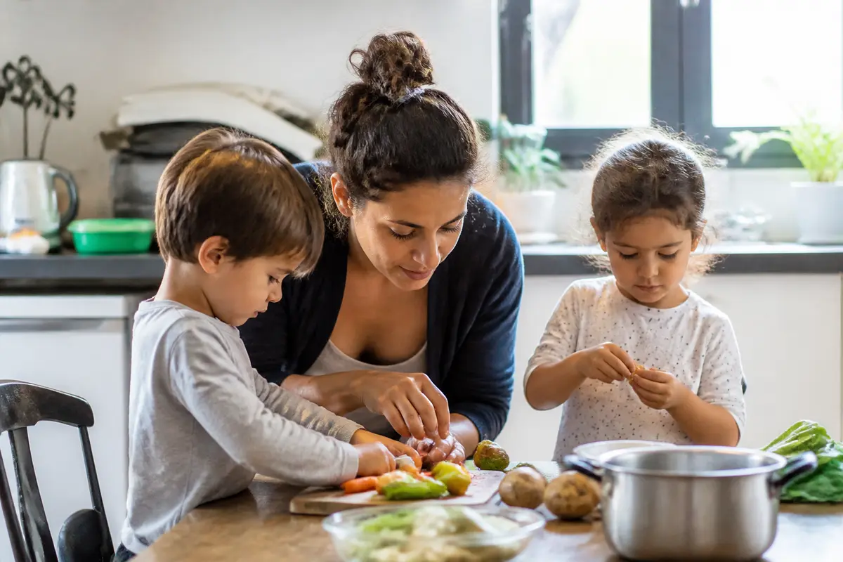 Une femme et deux enfants préparent des légumes ensemble dans une cuisine lumineuse.