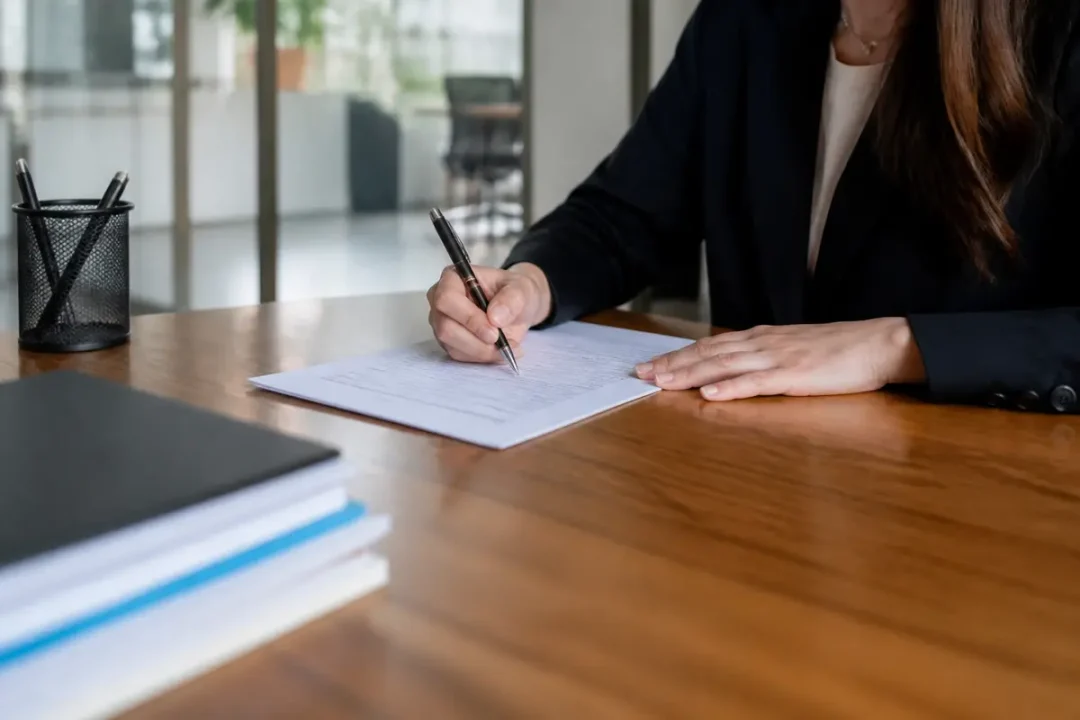 Une personne en costume noir signe des documents sur un bureau en bois, stylo à la main.