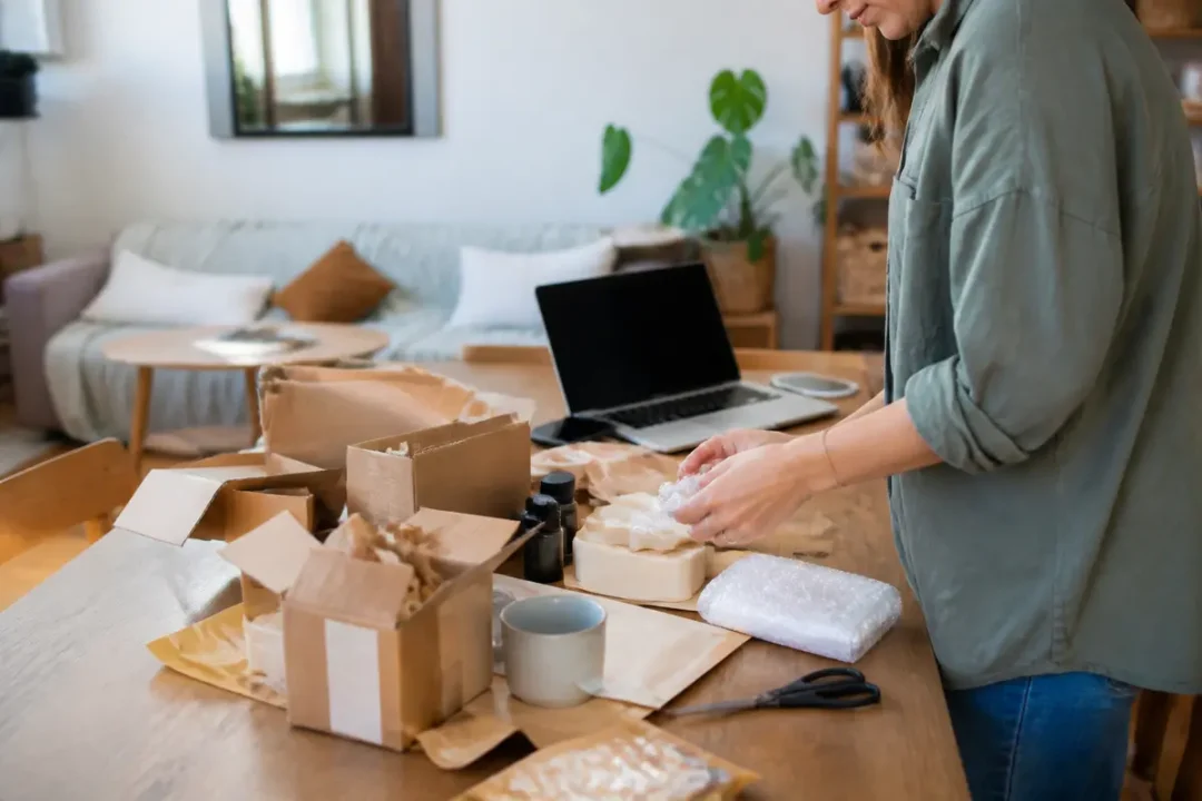 Personne emballant des objets avec papier bulle, cartons sur table, ordinateur portable en arrière-plan.